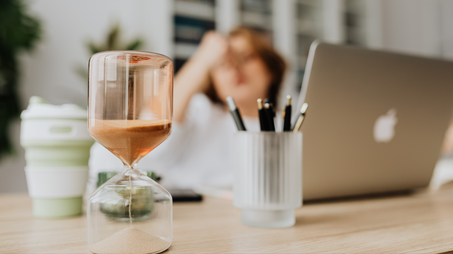 An hour glass sitting on a desk in front of someone holding the bridge of their nose sitting in front of their laptop looking stressed. 